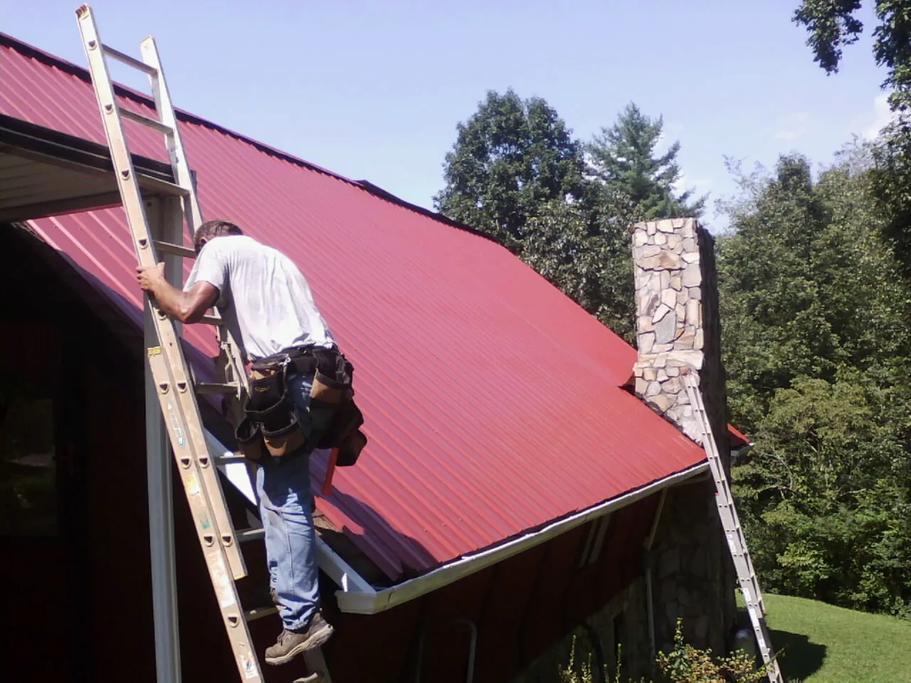 Roofer on a ladder beside a red metal roof and stone chimney — Soffit & Fascia Repair by Roofing Brothers in Woodhull